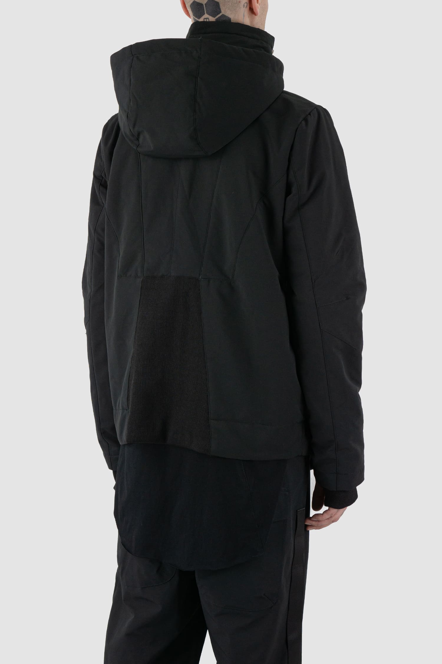 First Aid to the Injured - back side view of black padded jacket with double zipper closure for men from the FW24 collection with detachable hood, 2 long zippers to close the jacket and knitted-in cuffs with thumb holes.